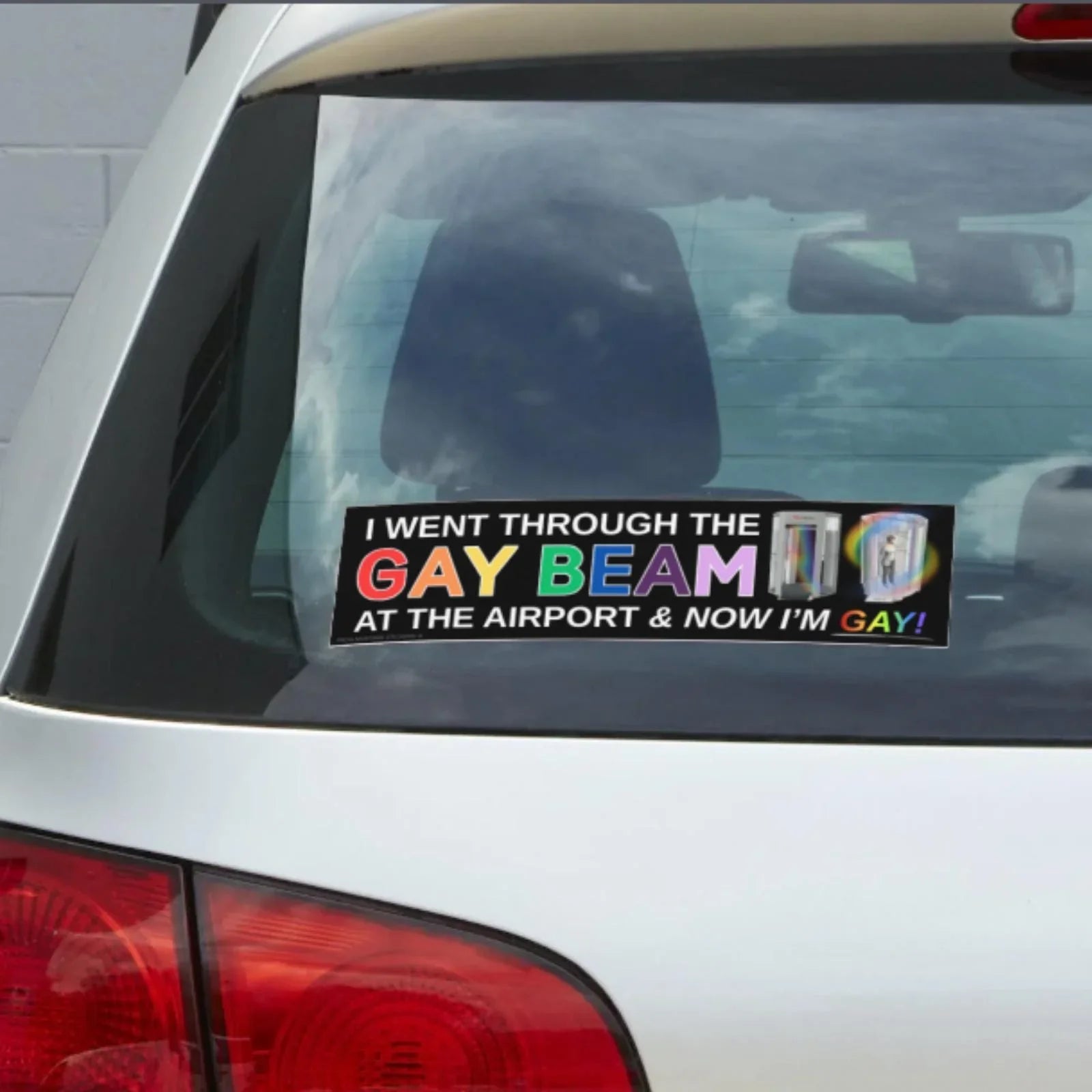 Funny rainbow text bumper sticker on car rear window, says 'I went through the gay beam at the airport & now I'm gay'.