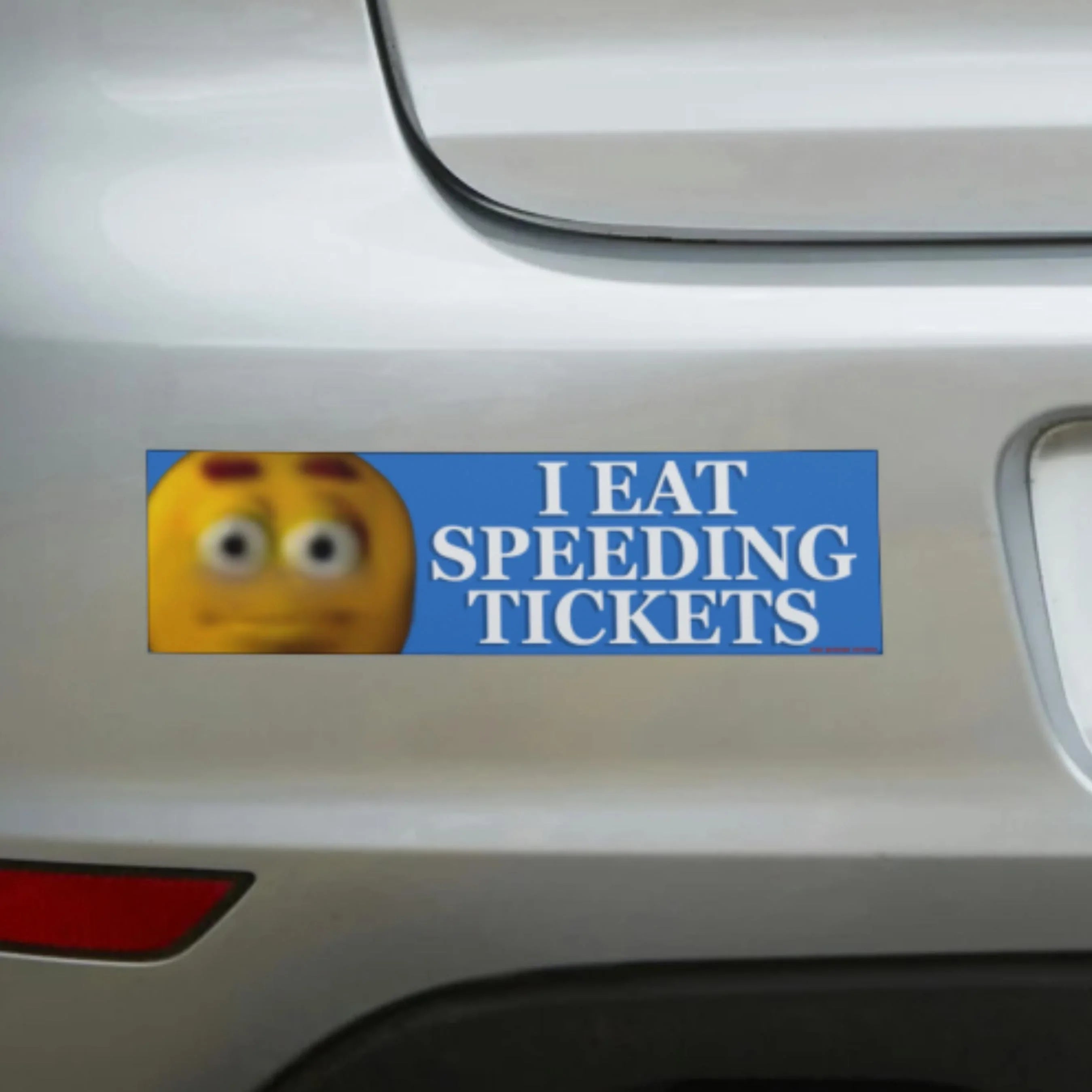 Funny bumper sticker saying 'I Eat Speeding Tickets' with emoji, on white car rear.